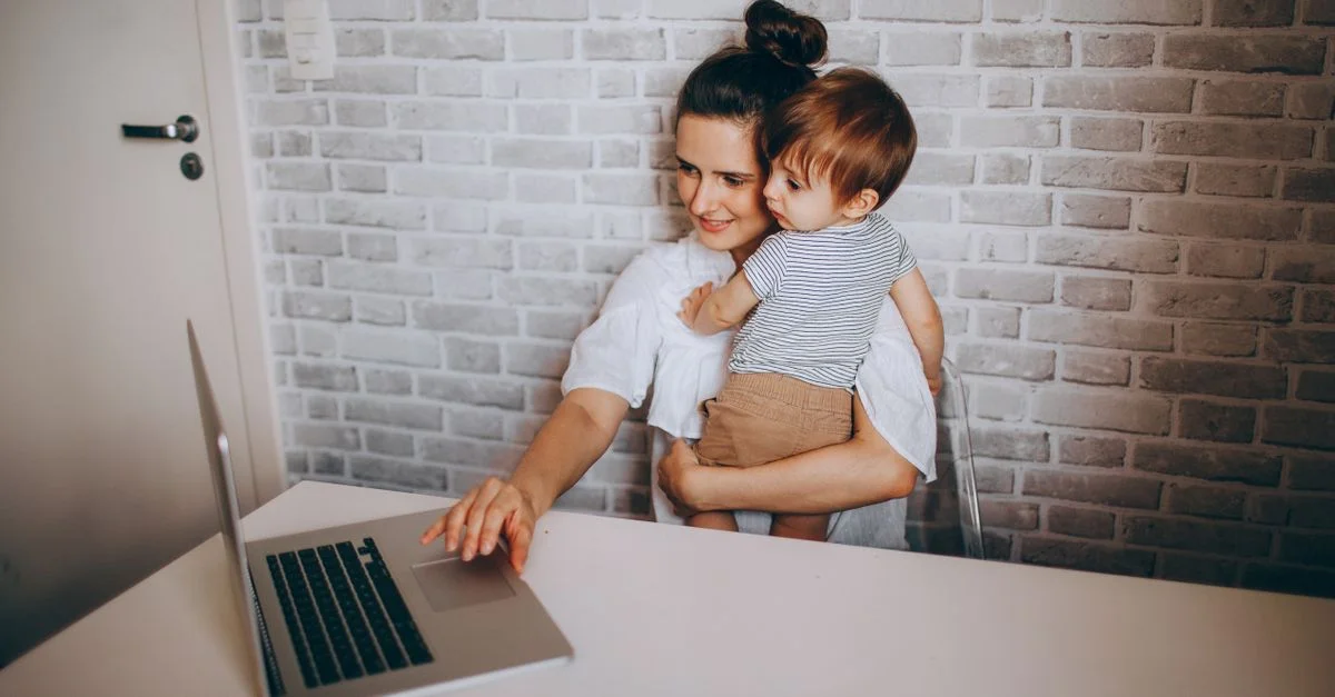 Une mère travaille à distance sur un ordinateur portable tout en tenant son tout-petit dans un cadre familial confortable.