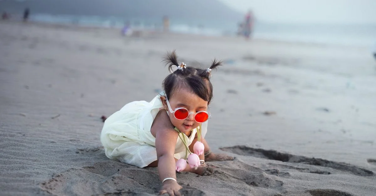 Une adorable petite fille profite d'une journée ensoleillée à la plage, portant des lunettes de soleil rouges et jouant dans le sable.