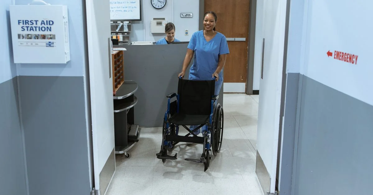 Une infirmière en blouse médicale pousse un fauteuil roulant vide dans un couloir d'hôpital.